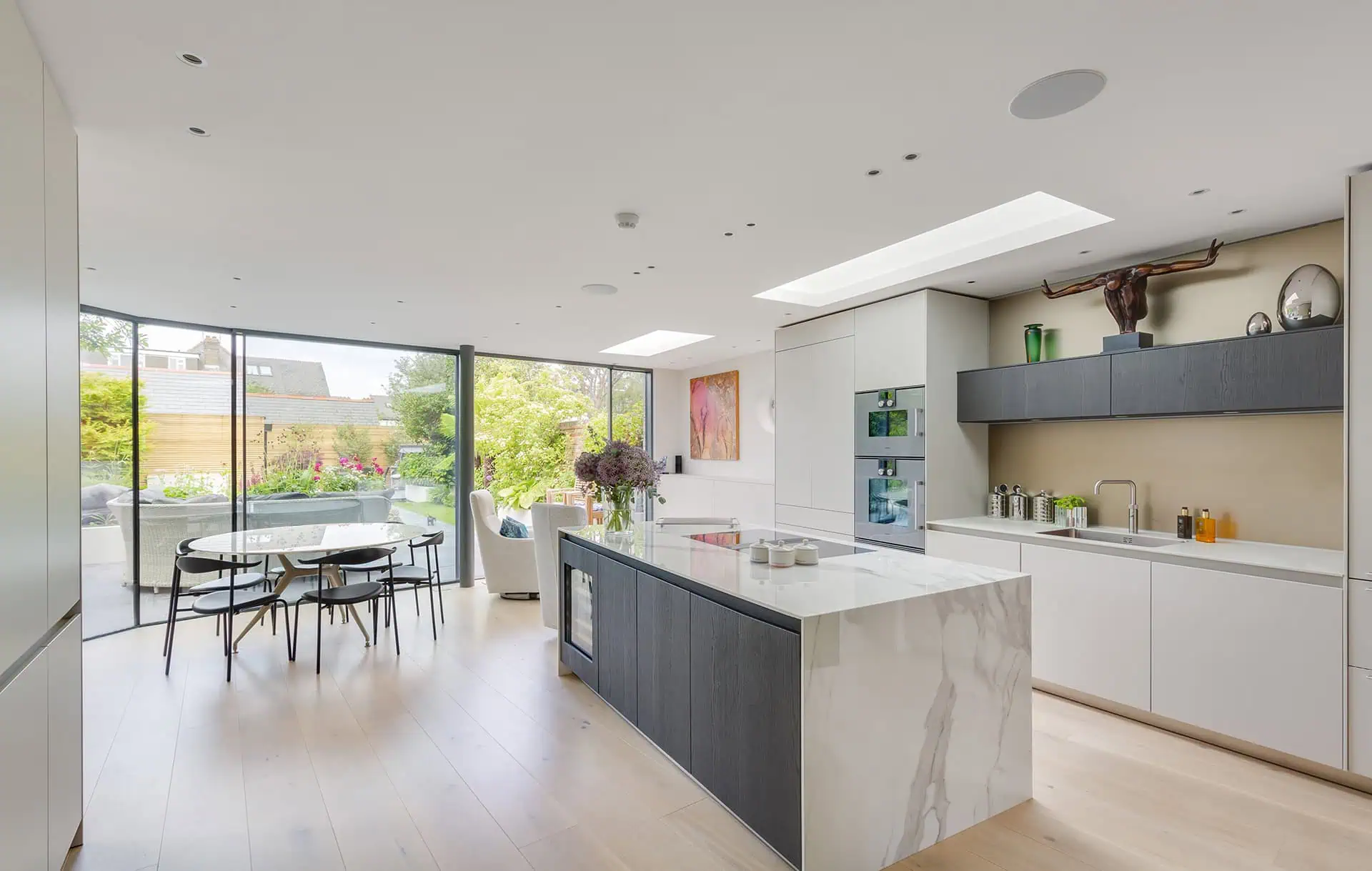 Modern kitchen interior in Belsize Park featuring a sleek island, minimalist cabinetry, and large glass windows opening to a lush garden, emphasizing luxury home refurbishment and design services.