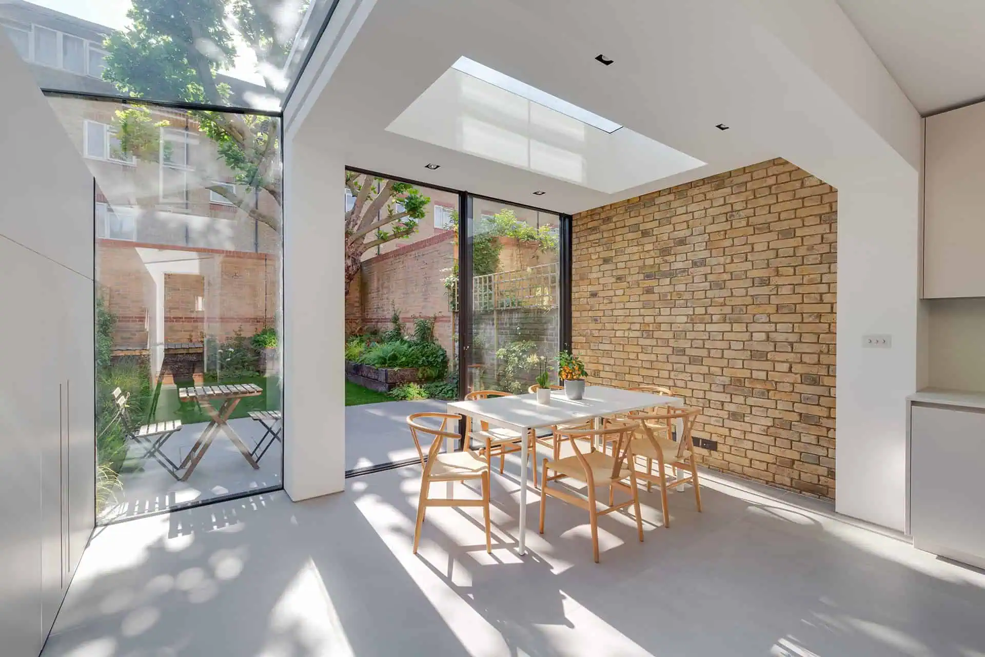 Modern dining area with wooden chairs and a white table, featuring large glass windows overlooking a lush garden, emphasizing luxury home refurbishment in Fulham.