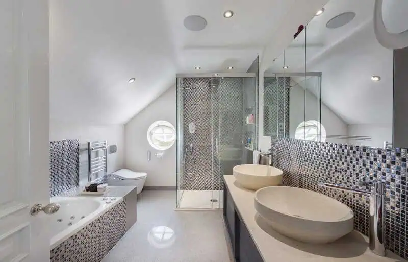 Modern bathroom design featuring a freestanding bathtub, dual vessel sinks, and a glass shower enclosure, showcasing stylish mosaic tile accents and natural light from a circular window, reflecting high-quality craftsmanship in Hampstead renovations.