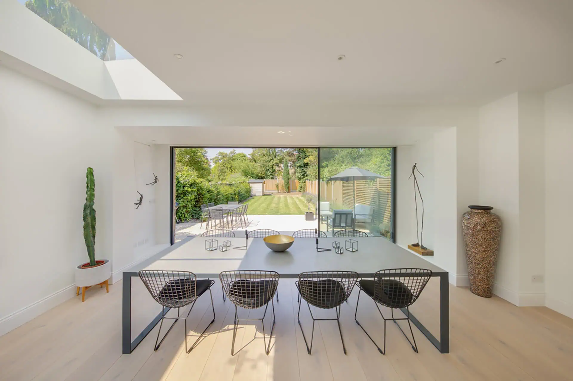 Modern dining area with large glass doors opening to a landscaped garden, featuring a sleek table, wire chairs, decorative bowl, and natural light enhancing the spacious design, reflecting luxury home extension in Kensington.